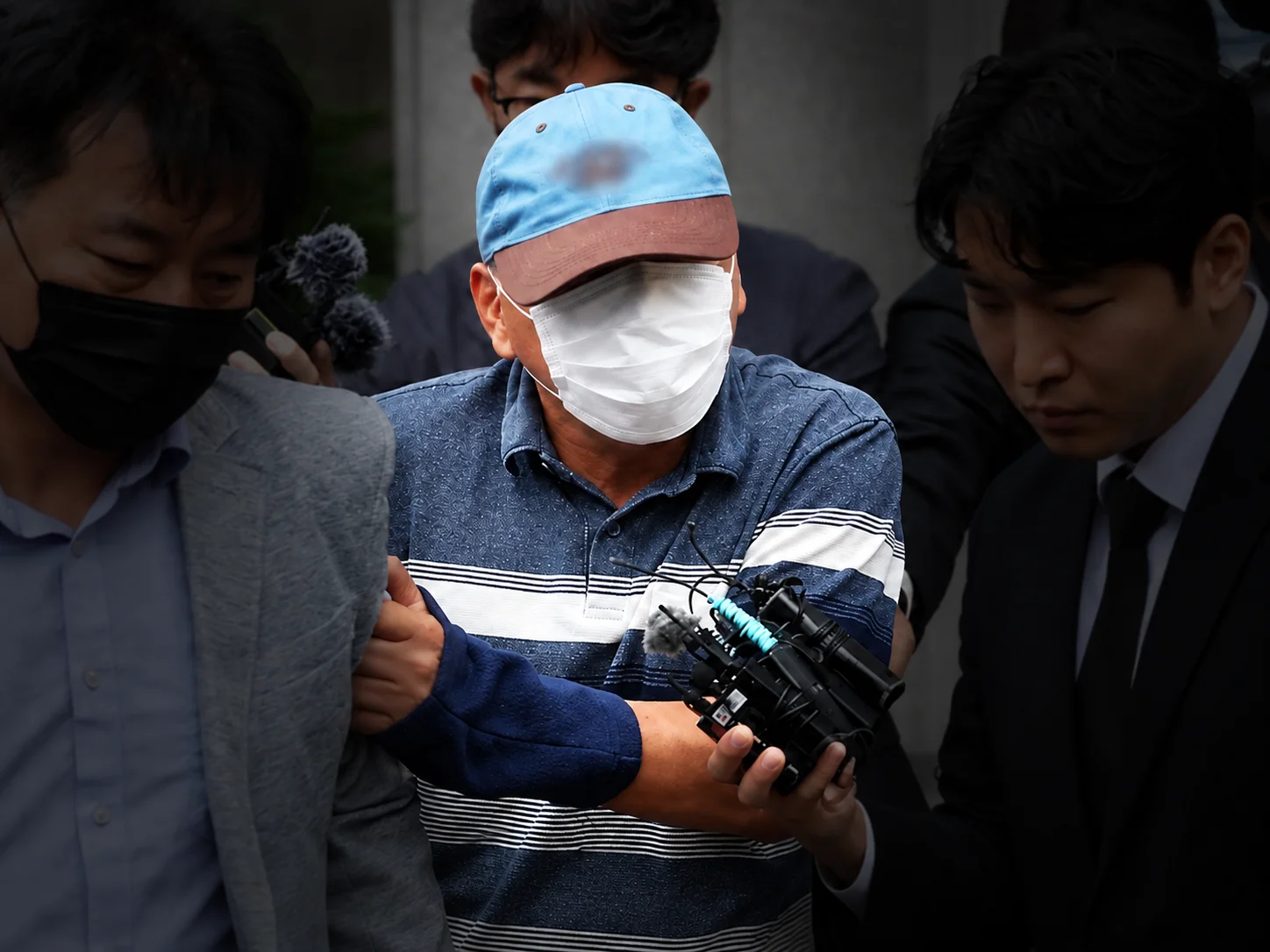 A man in a cap and face mask being escorted through a crowd of reporters