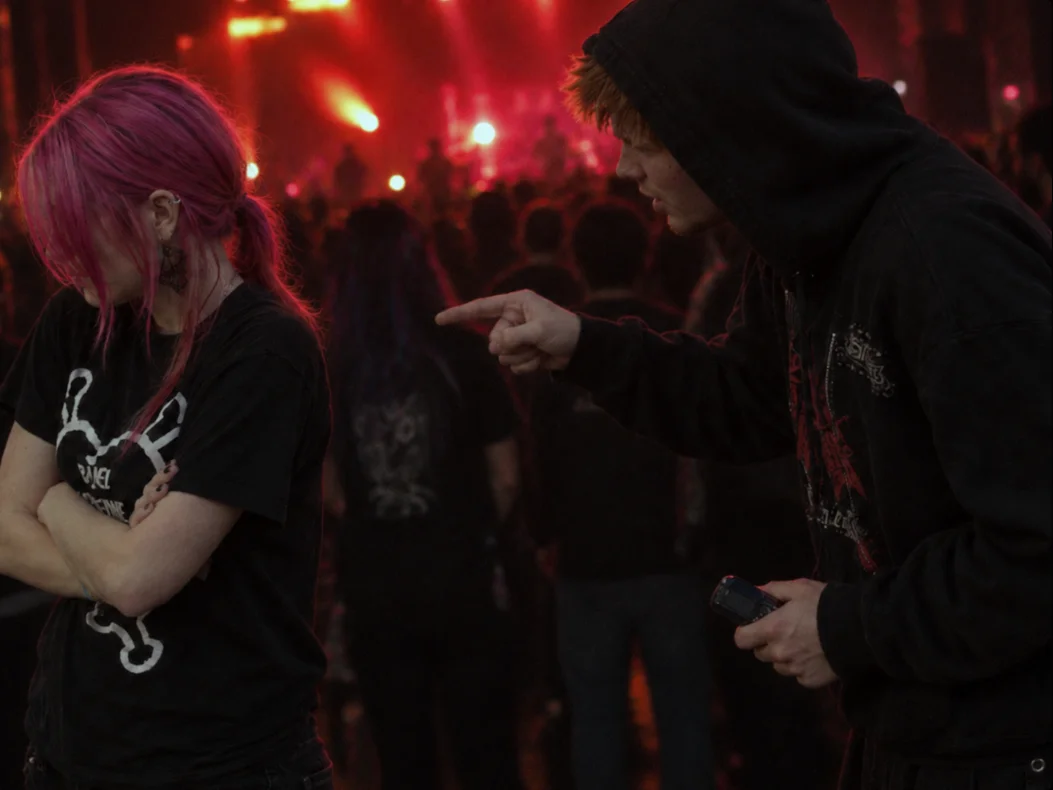 A hooded young man pointing at a girl with pink hair who stands with her arms crossed, a phone in his hand, in front of a concert crowd under red stage lights