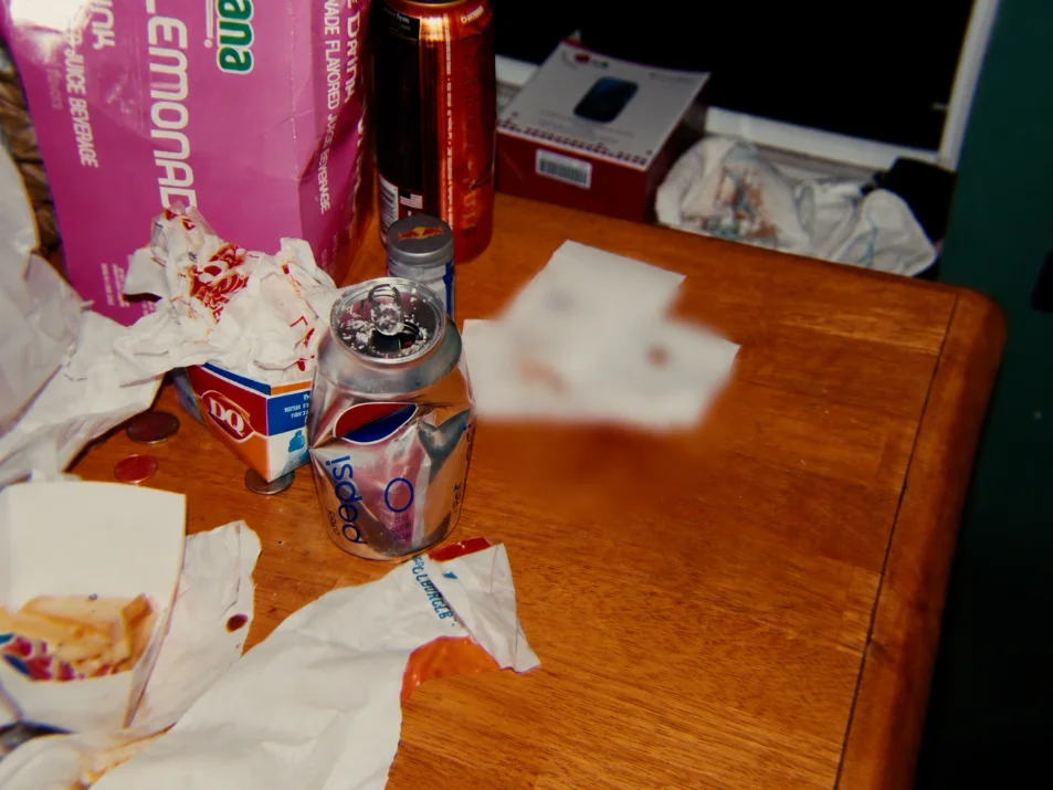 A wooden table cluttered with food wrappers, a fast-food container and a crushed soda can