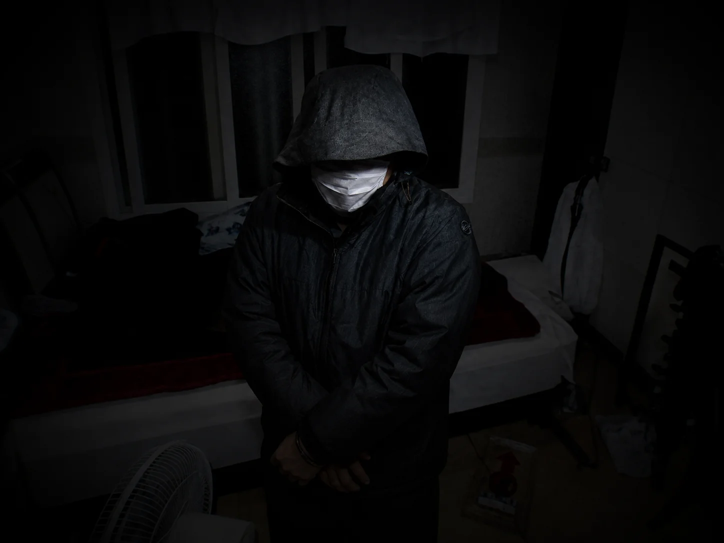 A hooded, masked man in handcuffs standing in a dark bedroom