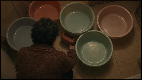 An older woman crouching among plastic basins on a tiled floor, with a red fuel can beside her