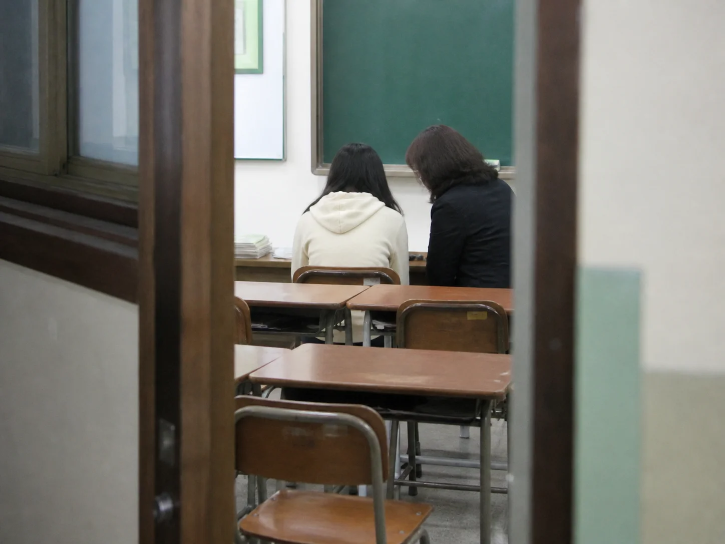 A student and a teacher sitting side by side in an empty classroom, seen from behind through the doorway