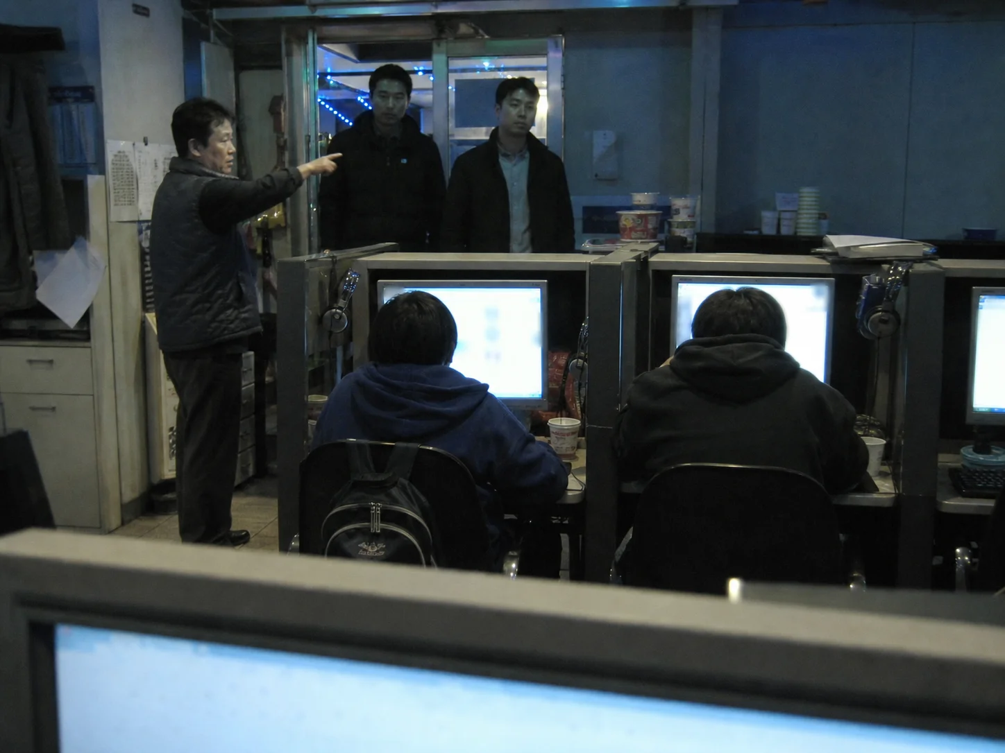 Men entering an internet cafe as one points toward two hooded figures seated at computers