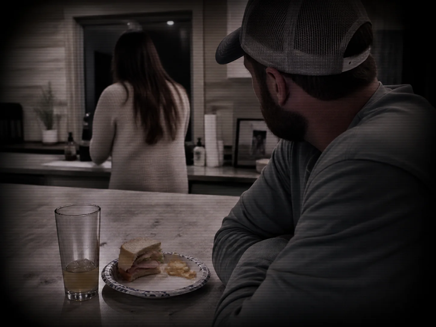 A man in a baseball cap glancing at a woman standing at a kitchen counter, with a half-eaten sandwich and a drink in front of him