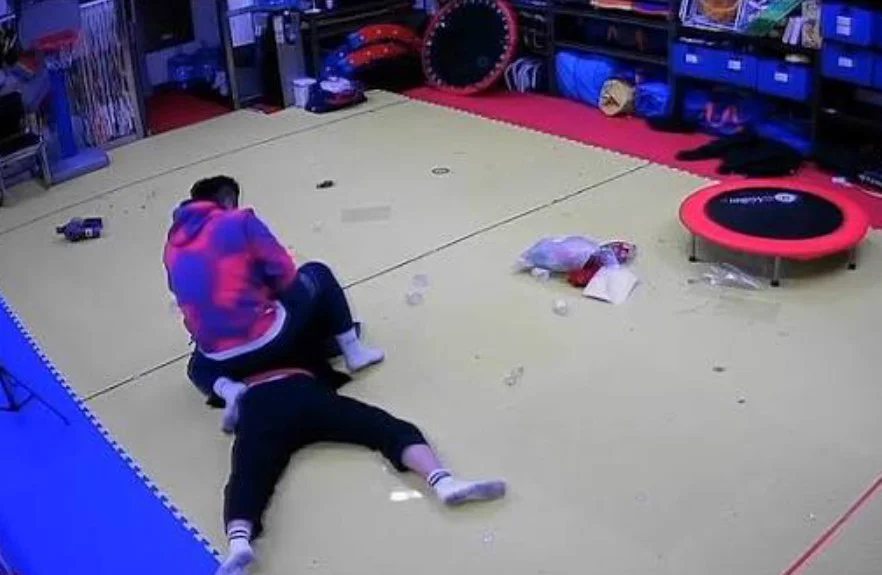 Security camera footage of a man in a pink and blue hoodie sitting on top of another man on a gym mat