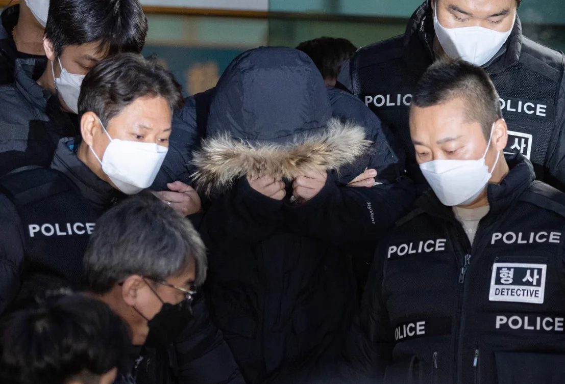 A man hiding his face under a fur-trimmed hood as detectives escort him through a crowd