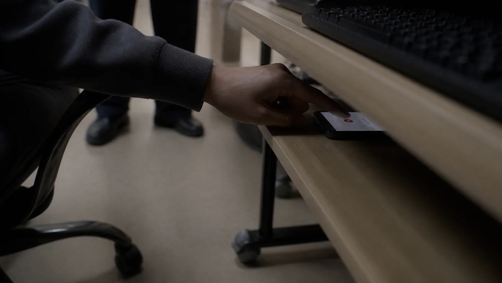 A hand pressing the record button on a phone tucked on a shelf under a desk