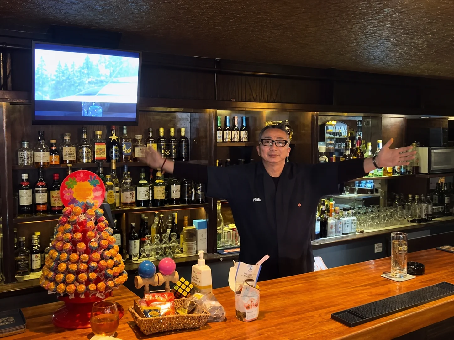 A man in glasses standing behind a bar counter with his arms spread wide, shelves of bottles behind him