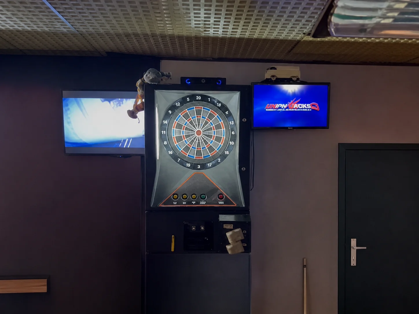 An electronic dart machine standing against a wall between two television screens