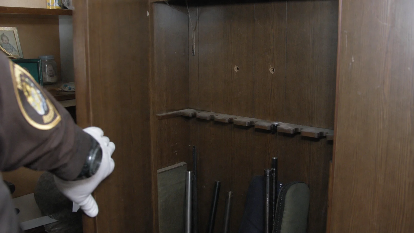 An officer's gloved hand beside an open wooden gun cabinet with two bullet holes in its back panel