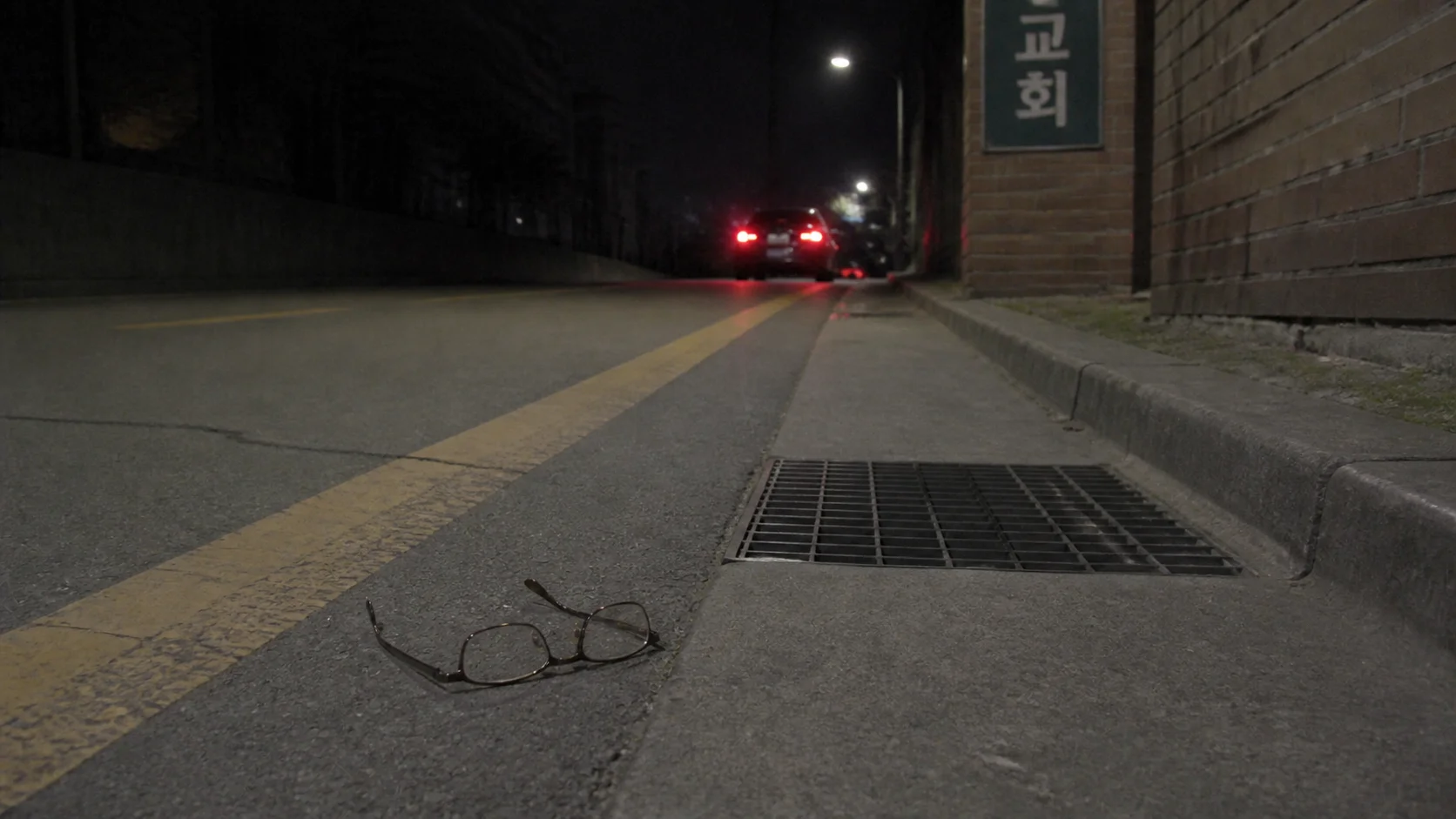 A pair of glasses lying on a road at night, with a car's tail lights in the distance