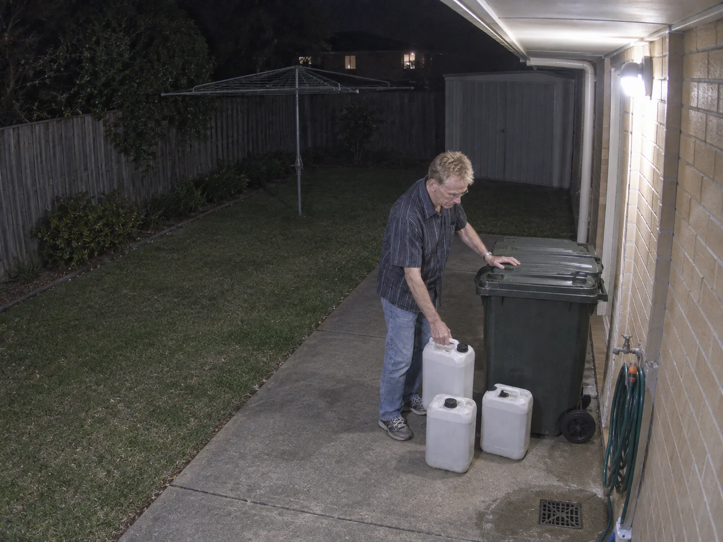 A man at night beside a wheeled trash bin with three white plastic containers on a concrete path next to a drain