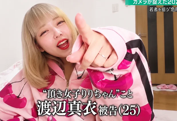 A blonde woman in a pink cherry-print jacket pointing at the camera, in a Japanese news broadcast captioned with the name Mai Watanabe, 25