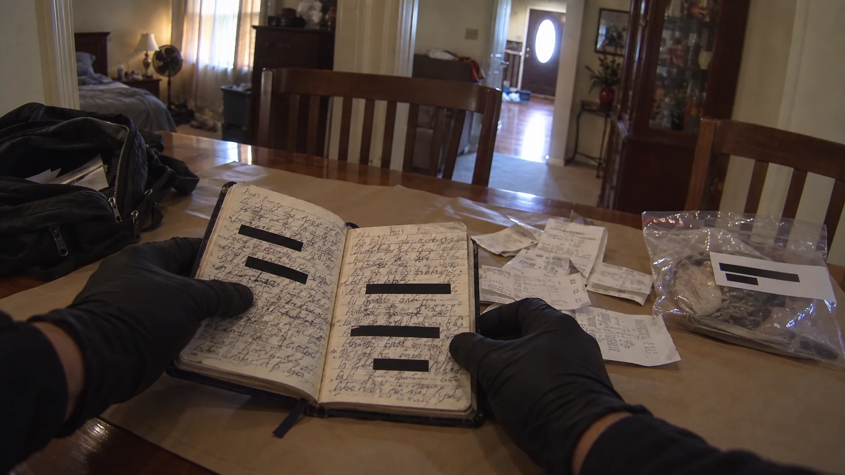 Gloved hands holding open a handwritten notebook on a table, with store receipts, an evidence bag and a black backpack beside it