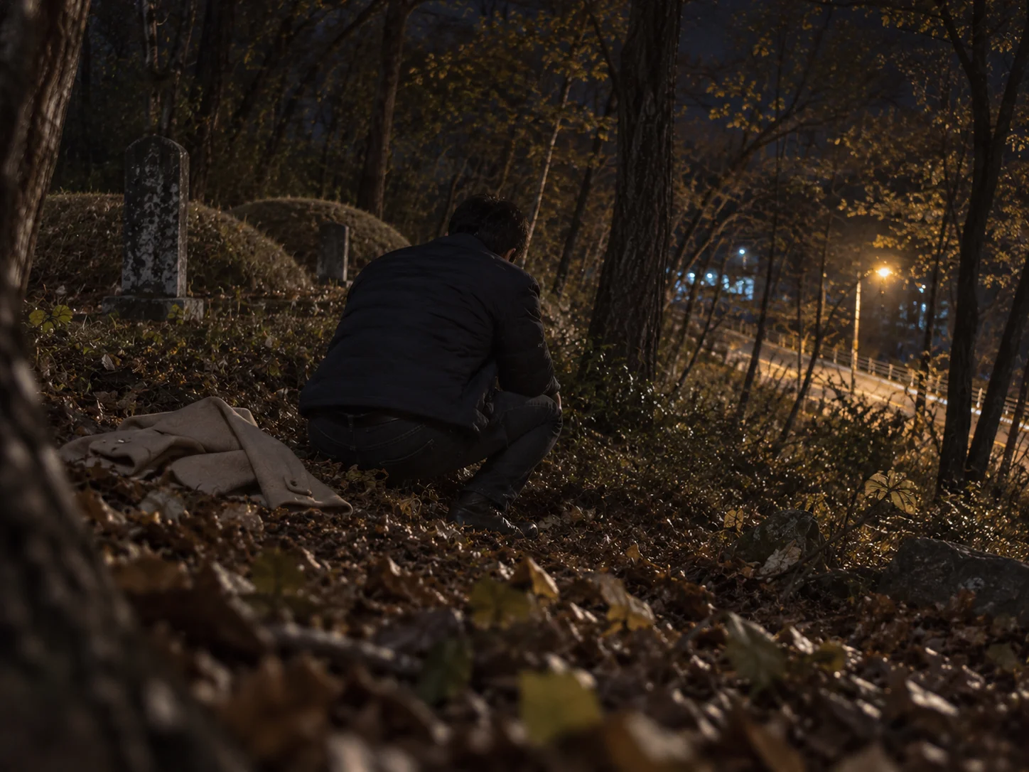 A man crouching at night on a leaf-covered wooded slope beside burial mounds and headstones, with a coat lying on the ground and a lit road below