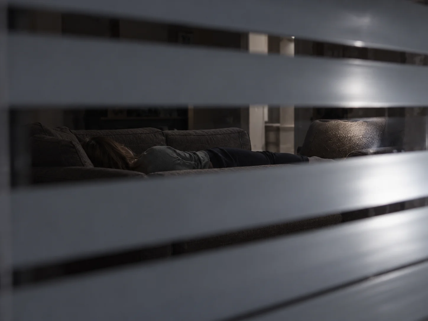 A view through the slats of window blinds into a dark living room, where a person lies face down on a sofa