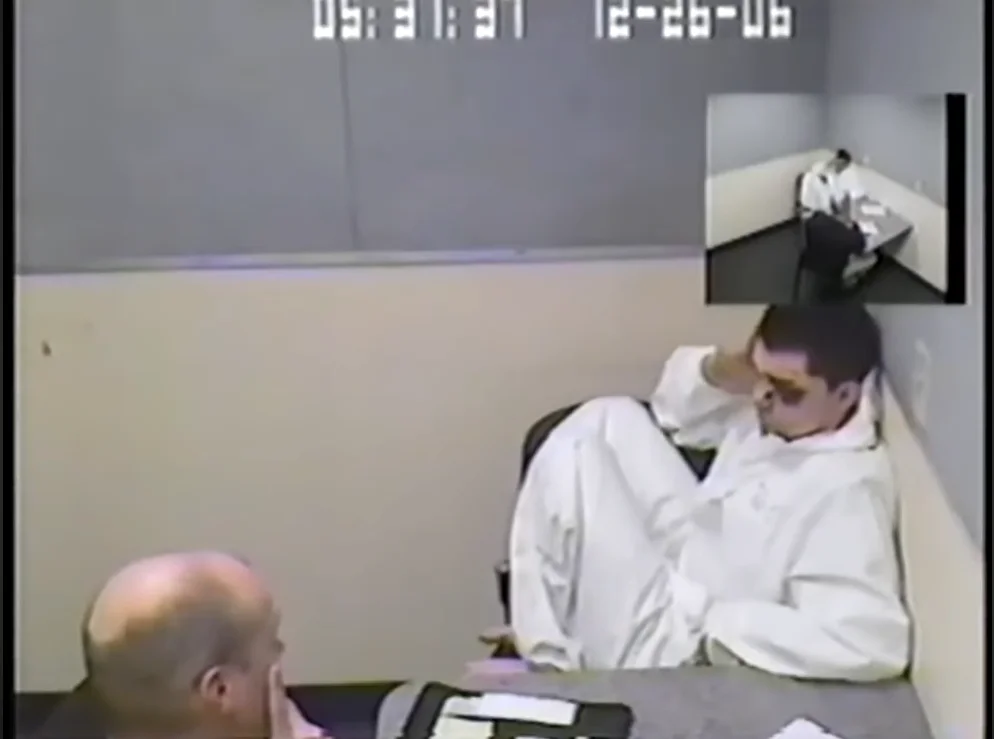 Interrogation room video timestamped 05:31 on 12-26-06, showing a young man in a white evidence suit slumped in a chair holding his head, with a detective in the foreground
