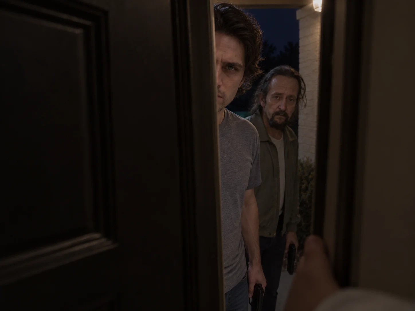 Two men holding handguns, seen through a partly open front door at night, with a hand pressing the door from inside