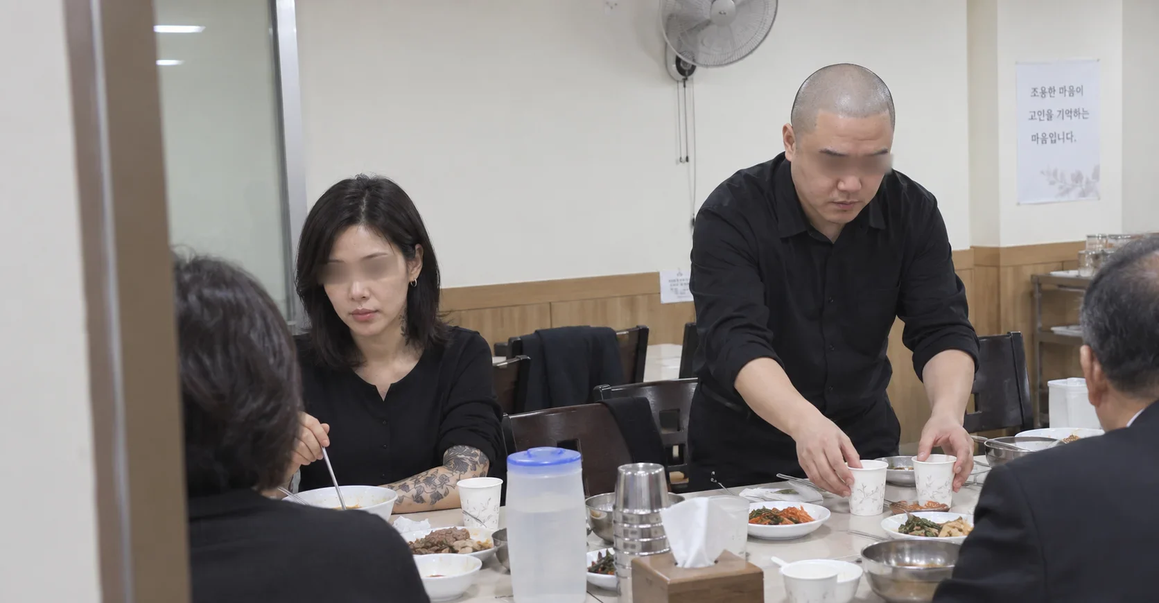 A shaved-head man in black setting out cups for mourners at a funeral hall dining table, beside a woman with a tattooed forearm