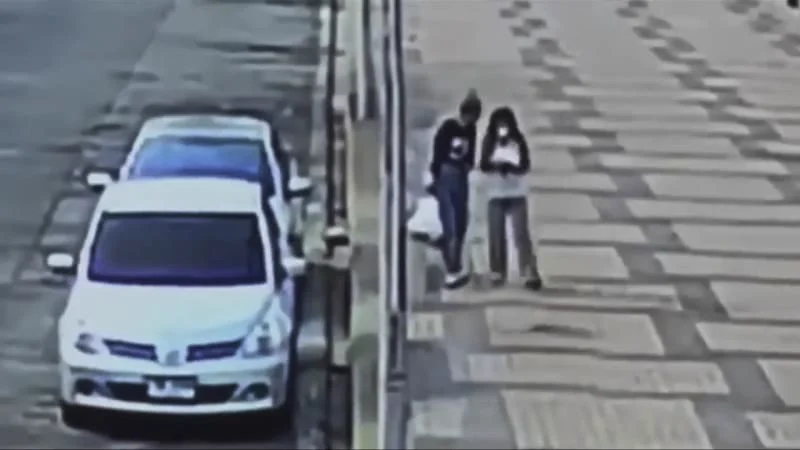 Grainy security camera footage of two women standing together on a wide sidewalk beside a parked white car
