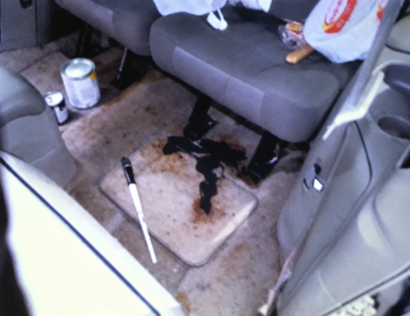 The rear floor of a car, where black paint has spilled across a reddish-brown stain on a floor mat, with paint cans nearby