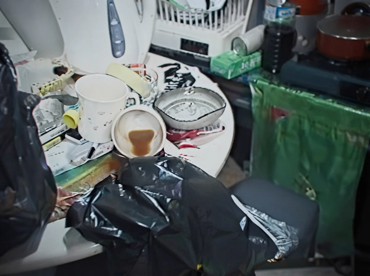 Two coffee cups, one with coffee left in it, on a cluttered kitchen counter beside a kettle and dish rack