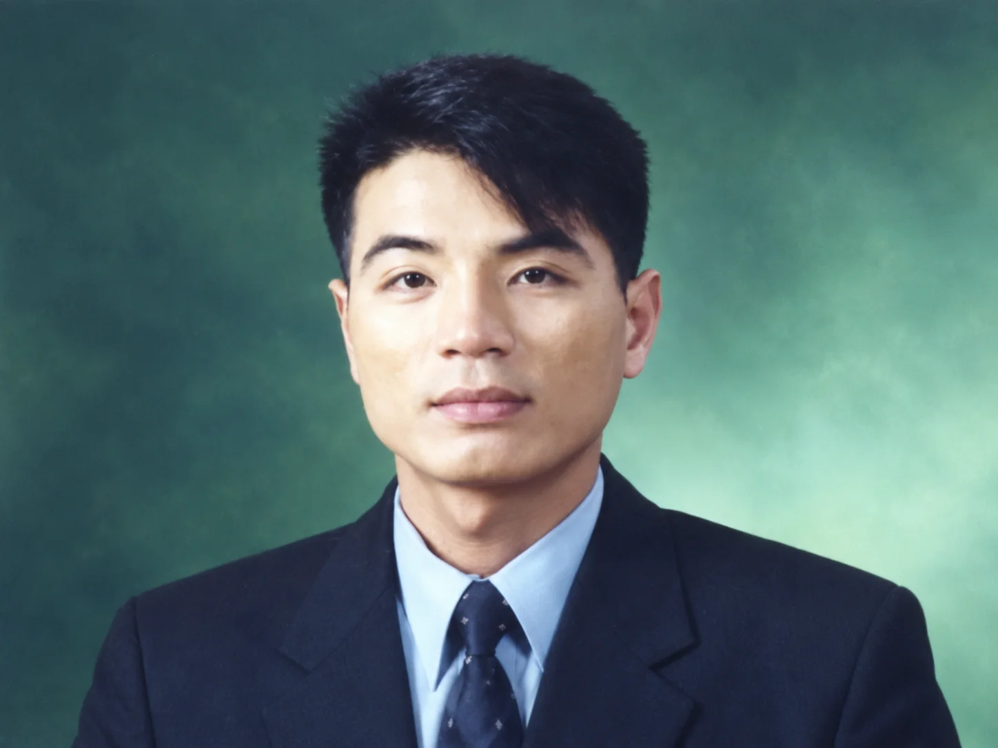Studio portrait of a man in a dark suit, light blue shirt and tie against a green background
