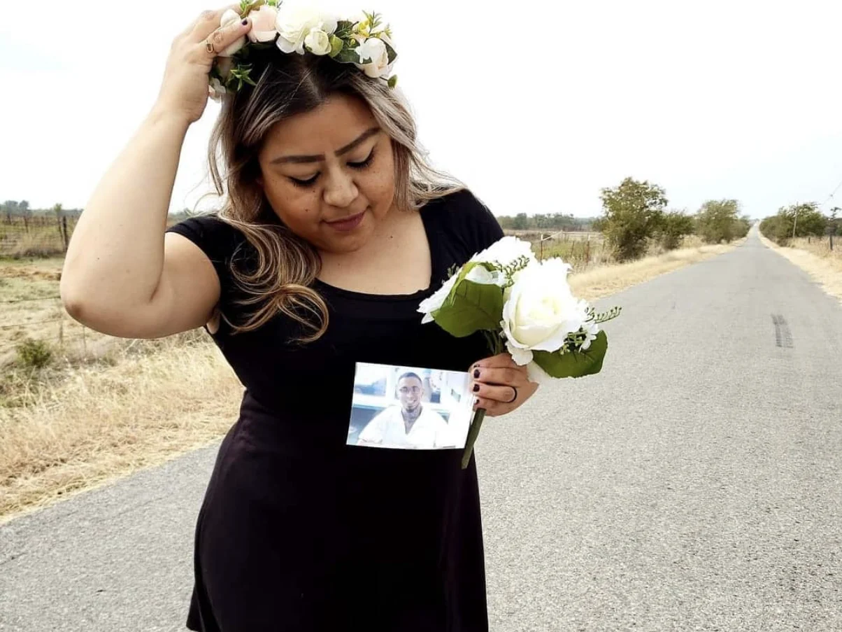 A woman in a black dress and flower crown standing alone on an empty country road, holding a bouquet and a photo of a man