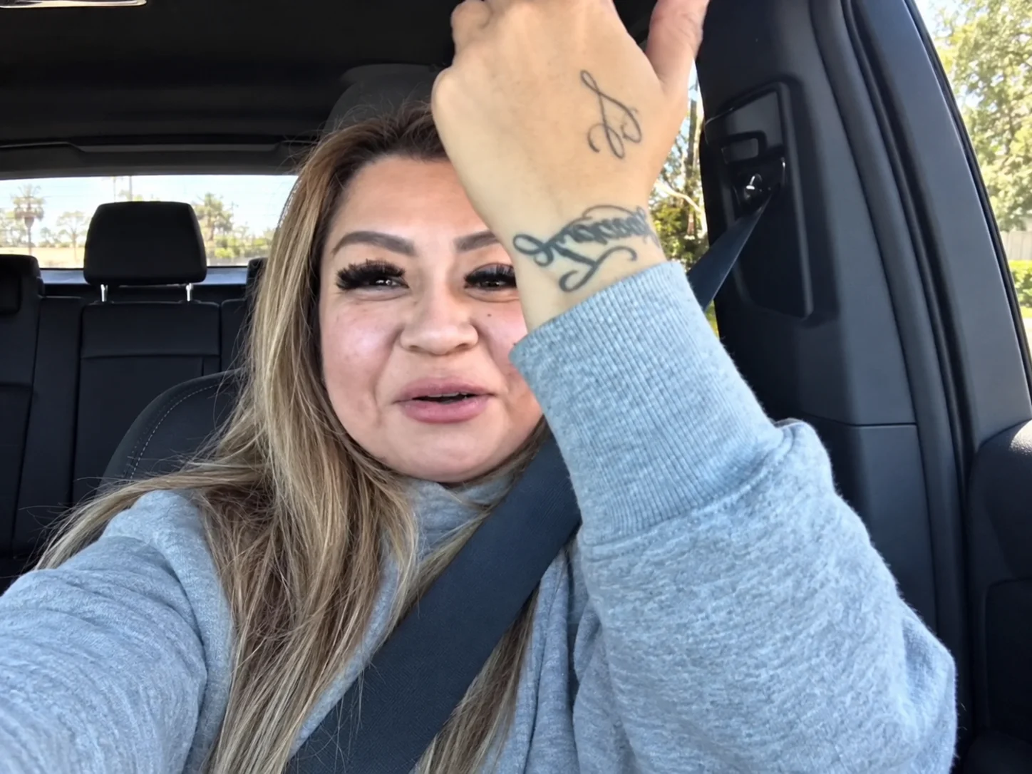 A woman filming herself in the driver's seat of a car, raising her hand to show a name tattooed on her wrist
