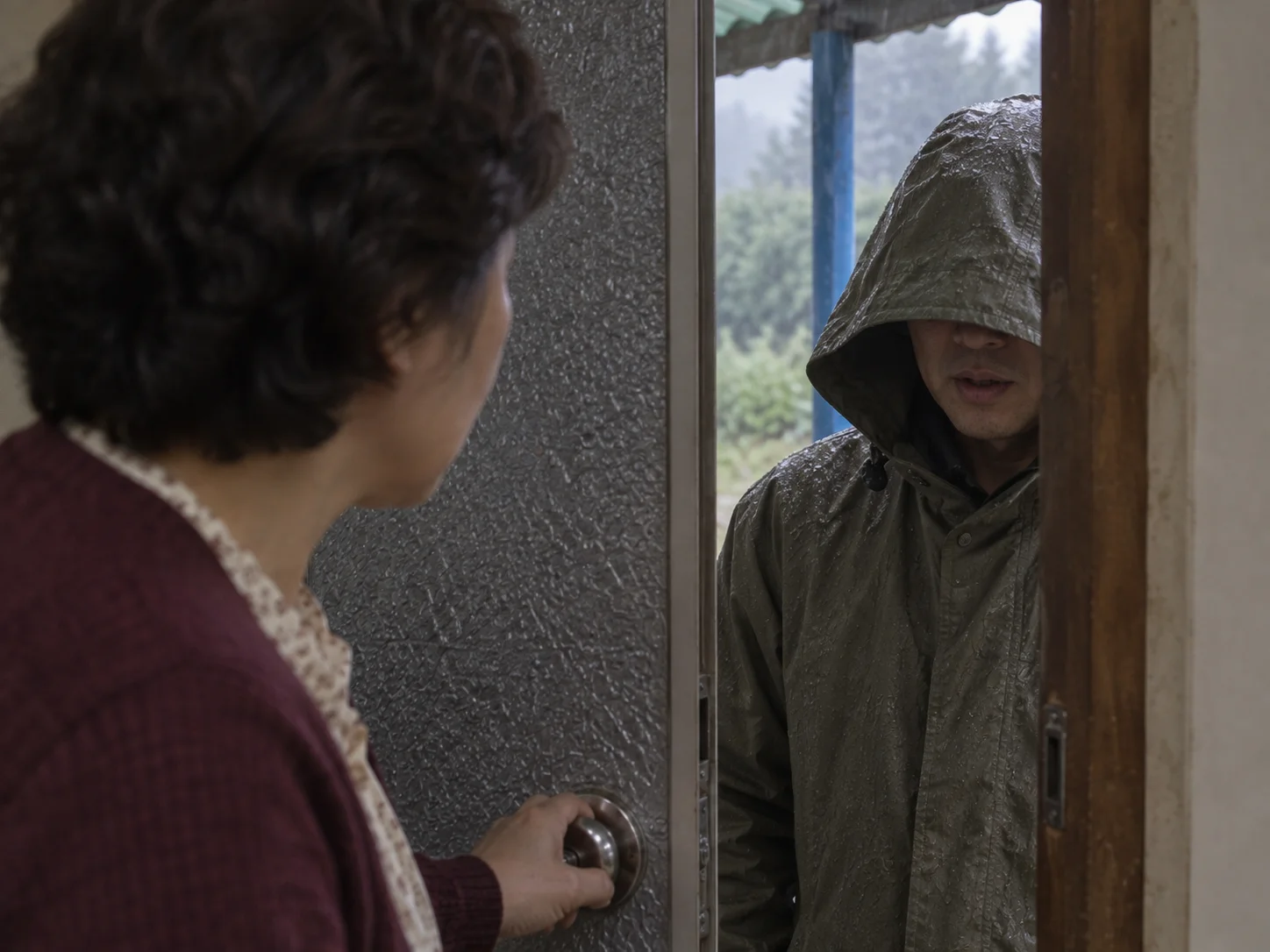 A woman holding a door open to a man in a wet, hooded khaki raincoat standing outside in the rain