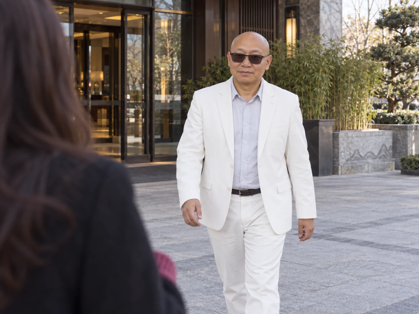 A bald middle-aged man in a white suit and sunglasses walking toward a woman outside a hotel entrance