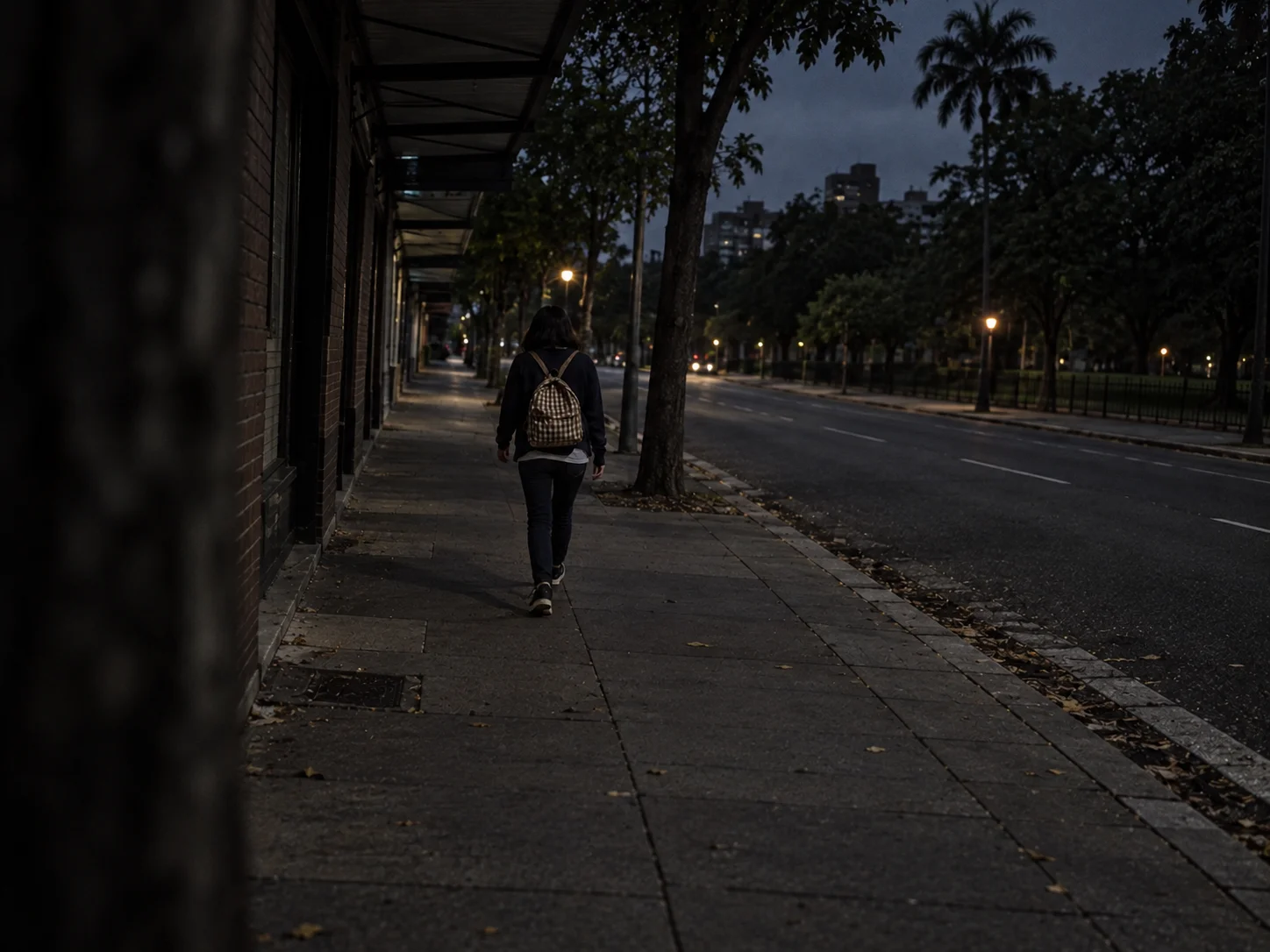 A young woman with a backpack walking alone along a dark city sidewalk before dawn